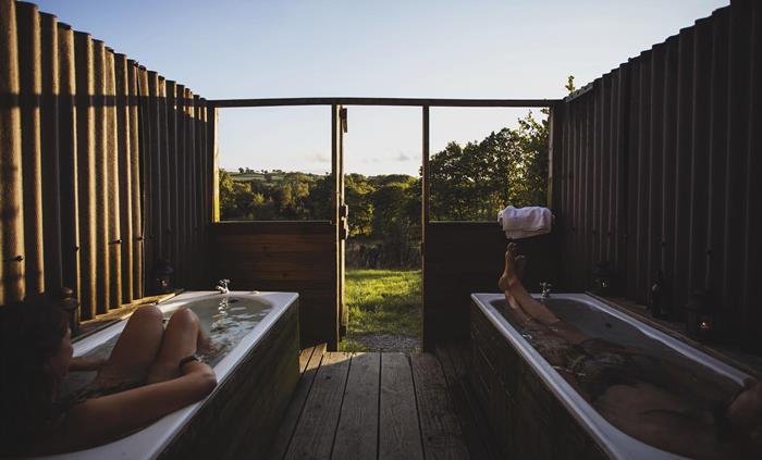 Two outdoor baths sit side by side with a view to the distant landscape of woodland and hills. The baths are full and there is a person soaking in eac